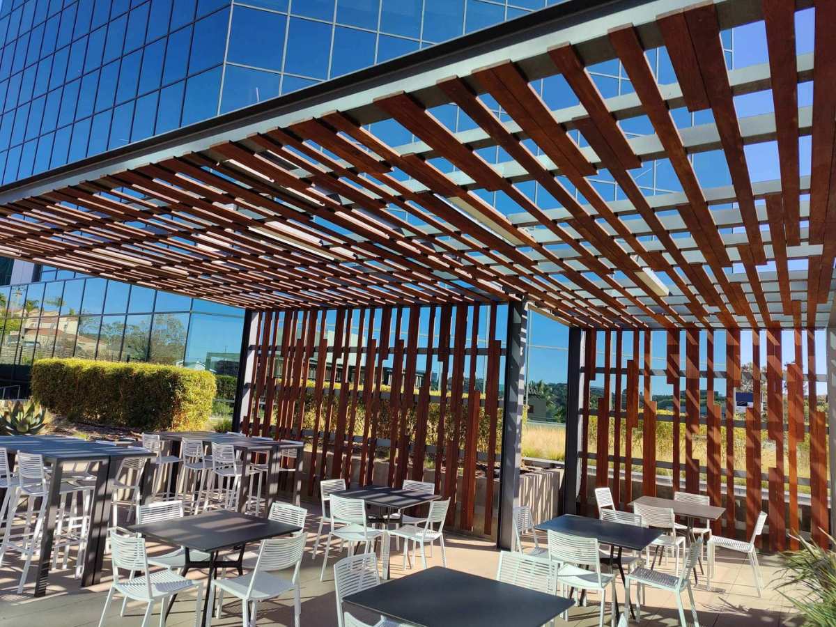 Outdoor dining area with modern wooden trellis, glass building reflections, and white tables and chairs in a sunny urban setting.