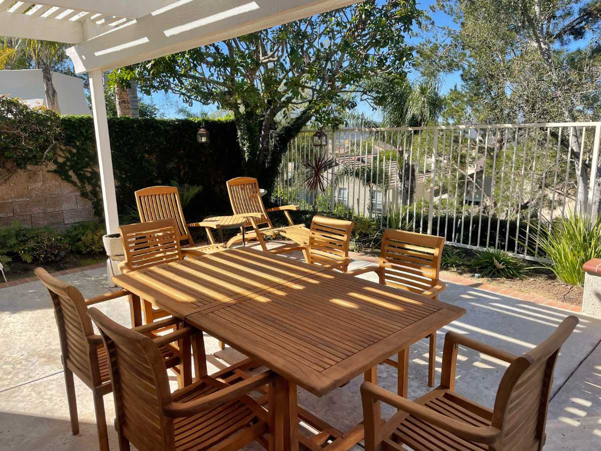 A sunlit outdoor patio featuring a wooden dining table and chairs, surrounded by lush greenery and a fenced area.