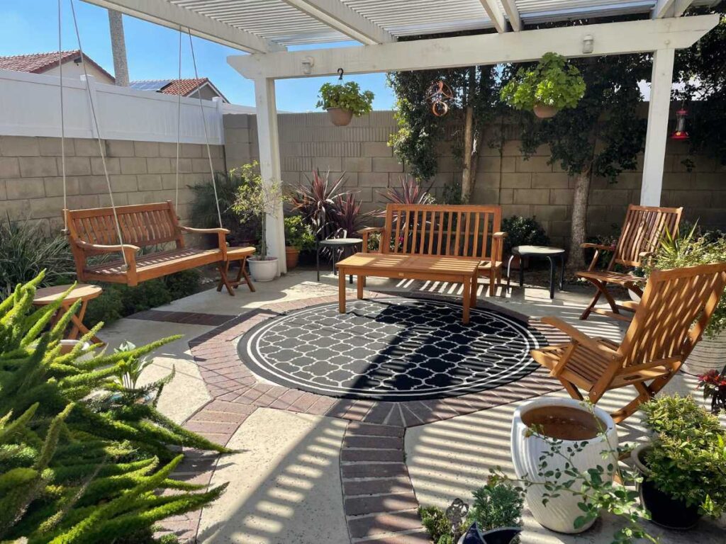 A cozy backyard patio with wooden furniture, a black and white rug, hanging plants, and surrounded by greenery under a shaded pergola.