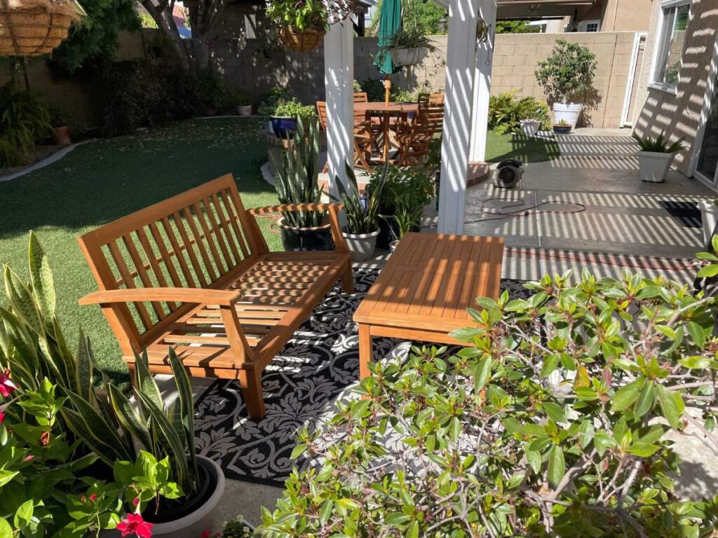 A cozy outdoor seating area with a wooden bench and table, surrounded by lush greenery and potted plants in a sunlit garden space.