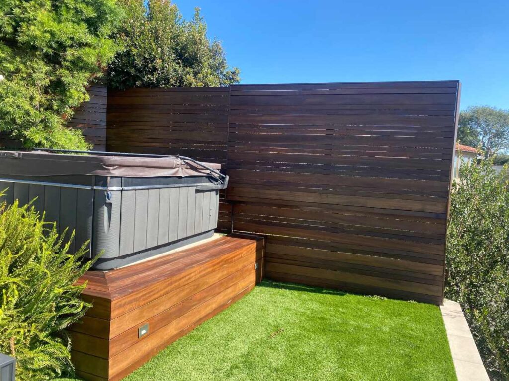 A hot tub sits on a wooden deck surrounded by lush greenery, with a slatted wooden fence in the background against a clear blue sky.