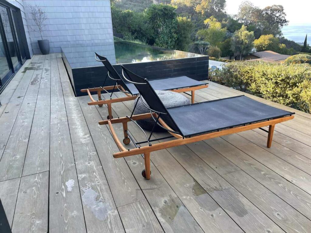 Two modern black loungers with wooden frames sit on a wooden deck, overlooking greenery and a serene pool in the background.