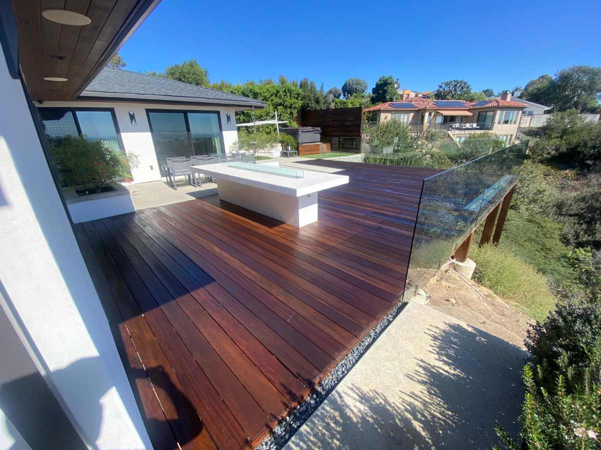 Modern deck with wooden flooring, glass railing, and a minimalist lounge area, surrounded by lush greenery and residential homes.