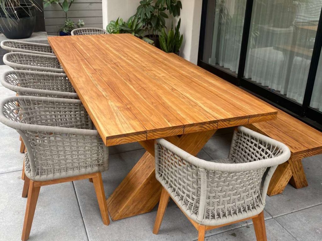 A modern outdoor dining set featuring a wooden table and six grey woven chairs on a tiled patio, surrounded by plants.