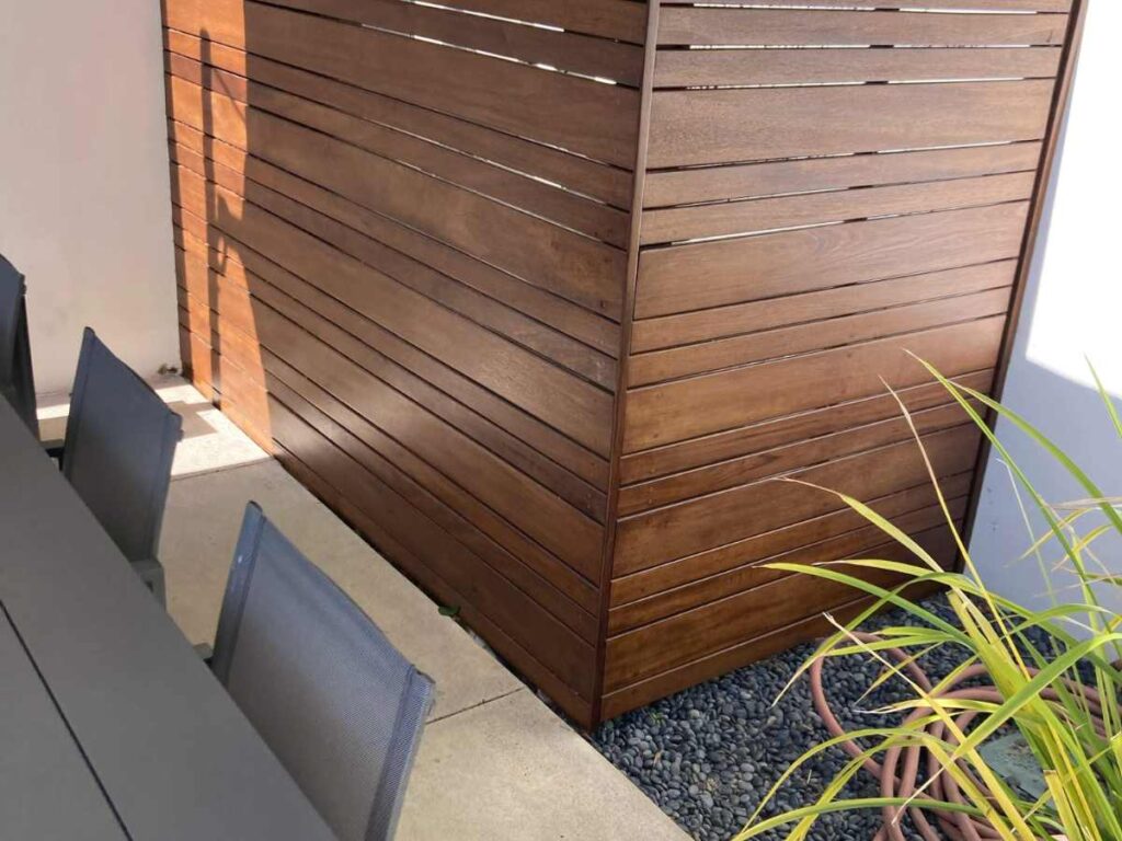 A modern outdoor dining area with a dark wooden privacy wall, grey chairs, and plants in the foreground, bathed in sunlight.