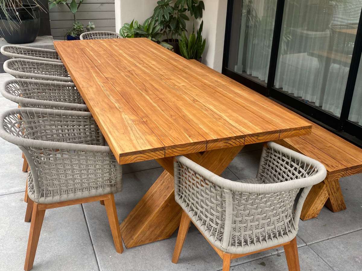 A modern outdoor dining set featuring a wooden table and six grey woven chairs on a tiled patio, surrounded by plants.