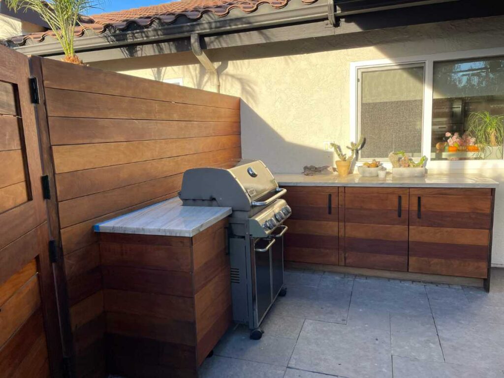 A modern outdoor kitchen featuring a stainless steel grill, wooden cabinetry, and a marble countertop, surrounded by lush greenery.