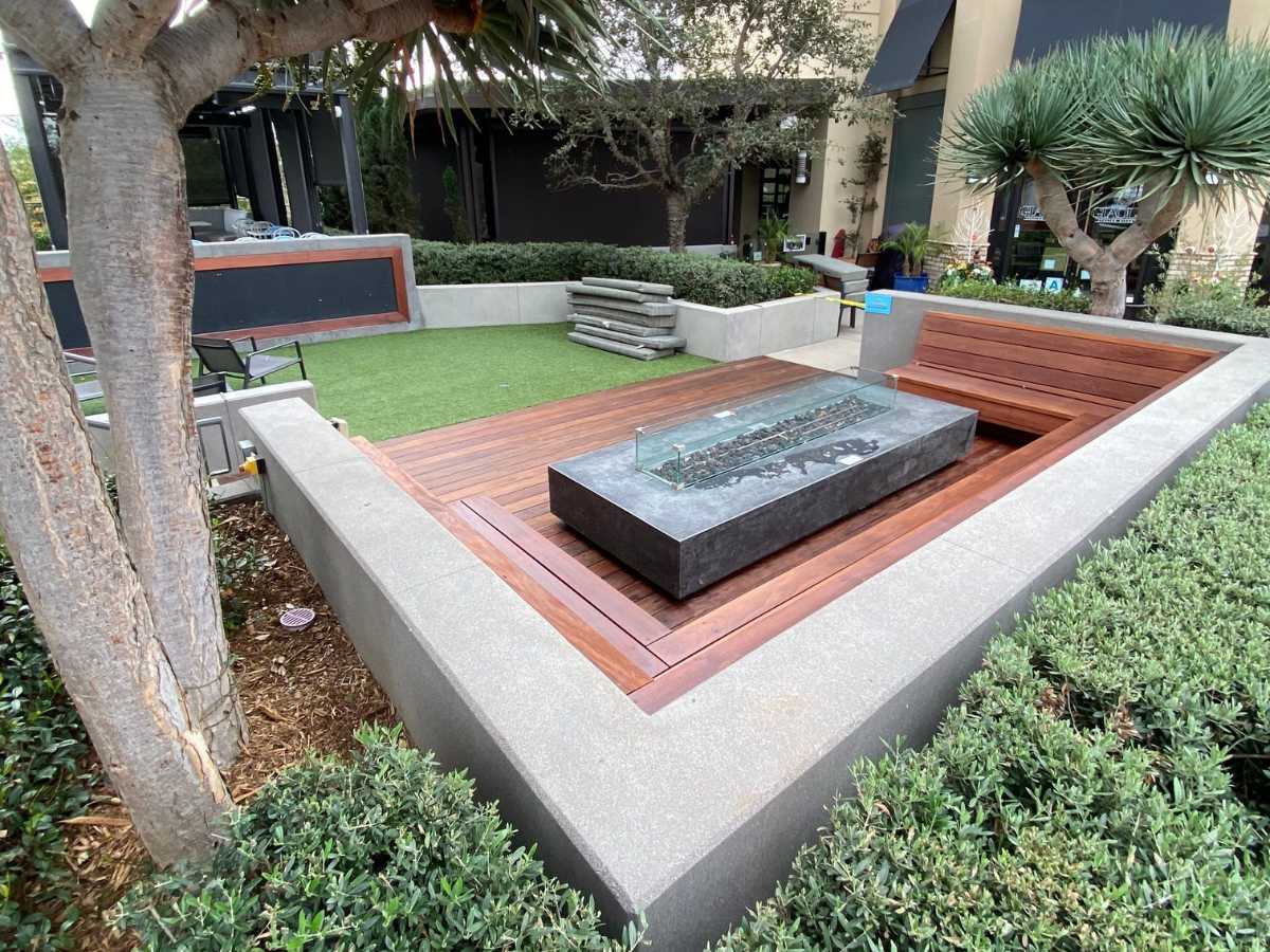 A modern outdoor lounge area featuring a wooden seating arrangement, stone table, and green grass surrounded by trees and shrubs.