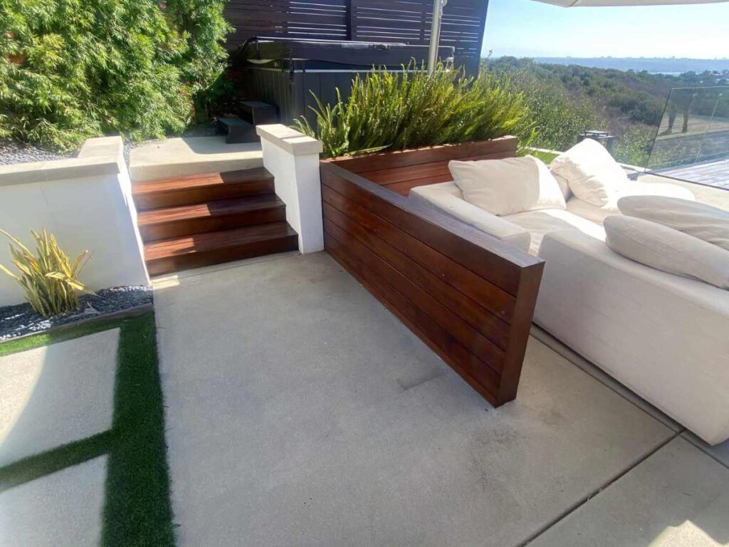 A modern outdoor patio features wooden seating, steps leading to a landscaped area, and lush greenery in a bright, sunny setting.