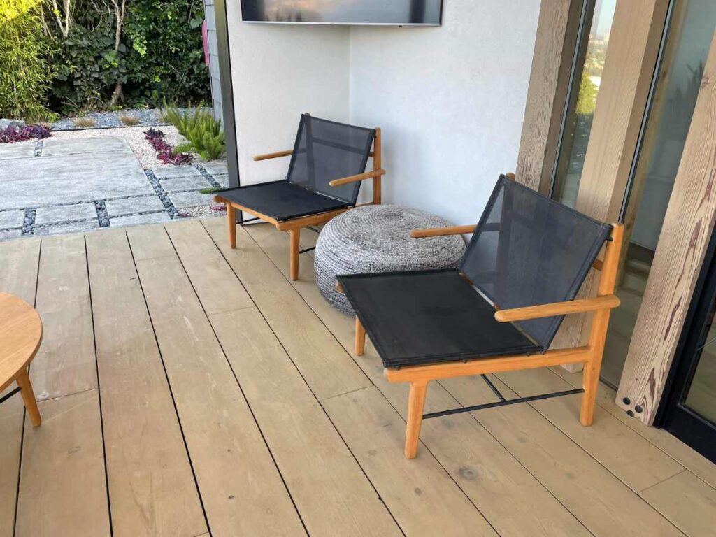 Two modern wooden chairs with black fabric seats are placed on a light wooden deck, next to a round cushion and a tv screen.