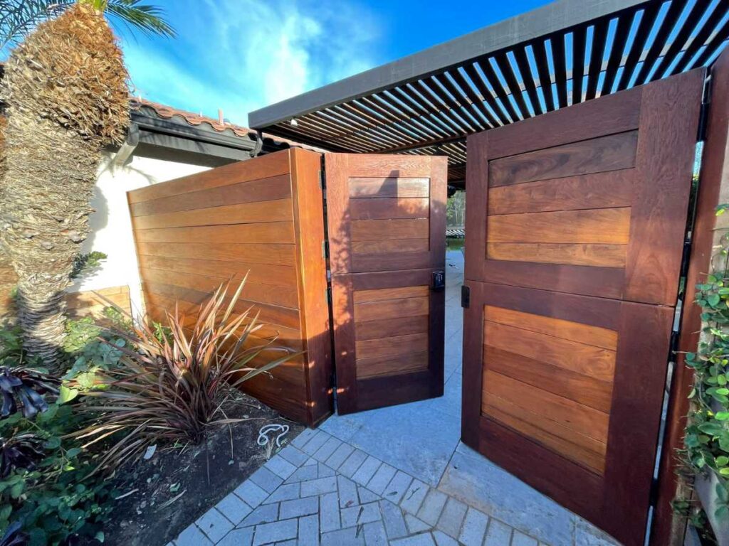 A wide view of a modern wooden gate opening into a landscaped area, flanked by lush plants under a bright blue sky.