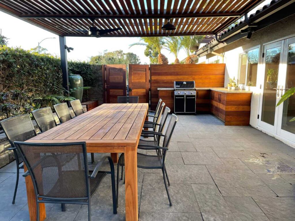 Outdoor patio featuring a large wooden dining table, metal chairs, and a built-in grill area under a slatted roof.