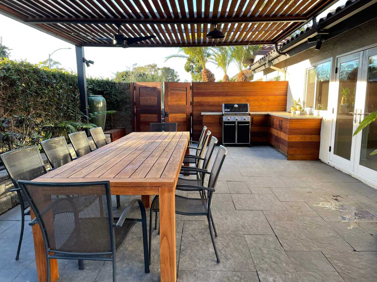 Outdoor patio featuring a large wooden dining table, metal chairs, and a built-in grill area under a slatted roof.