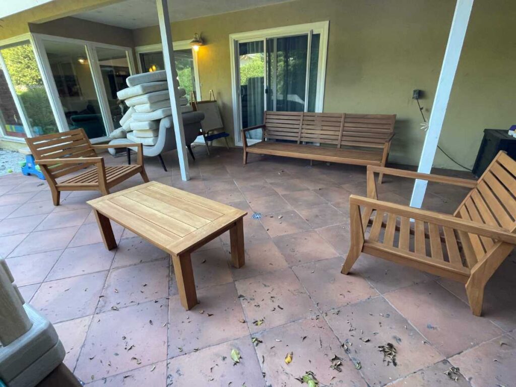 A patio with wooden furniture, including two armchairs and a table, beside a stack of cushions and a view into a living area.