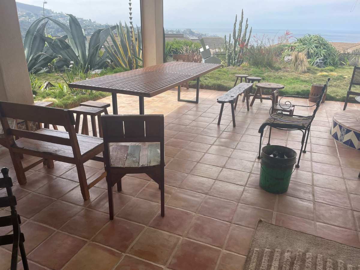A rustic outdoor seating area with wooden chairs and a large table, overlooking greenery and a distant seaside view under a cloudy sky.