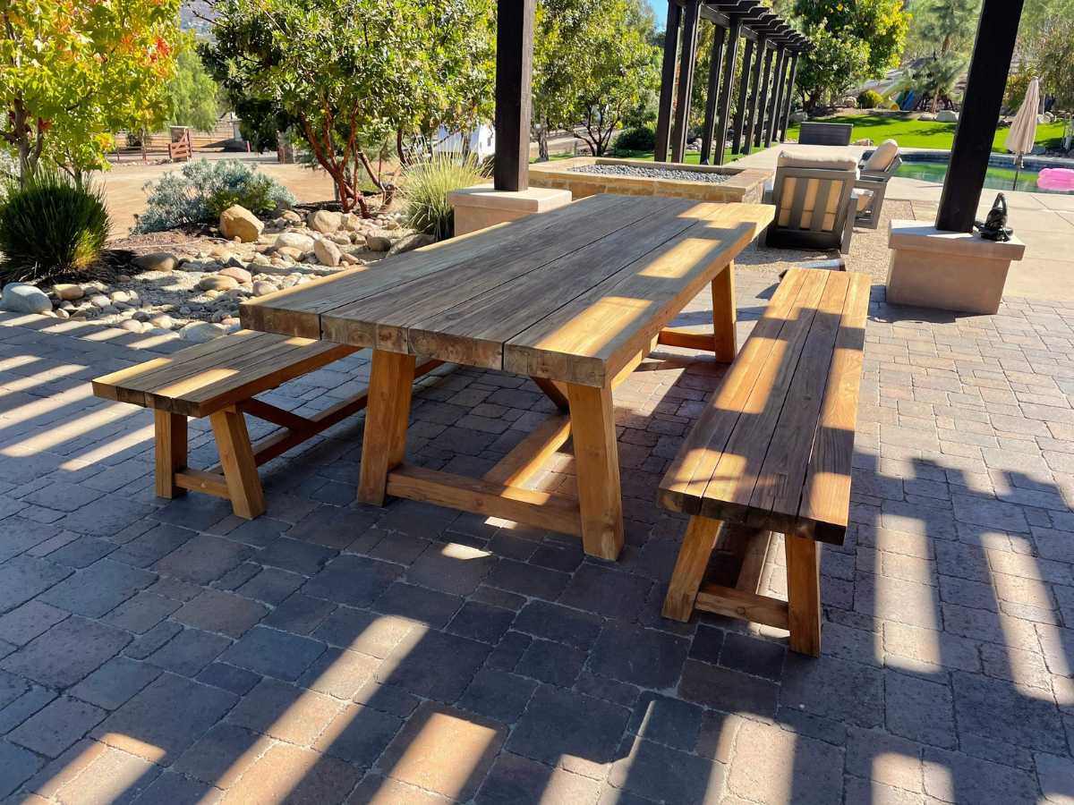 A rustic wooden picnic table with two benches sits on a stone patio, surrounded by greenery and shaded by overhead beams.