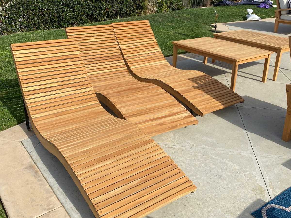 Three stylish wooden loungers with curved slats sit on a patio beside a simple wooden table, surrounded by grass and greenery.