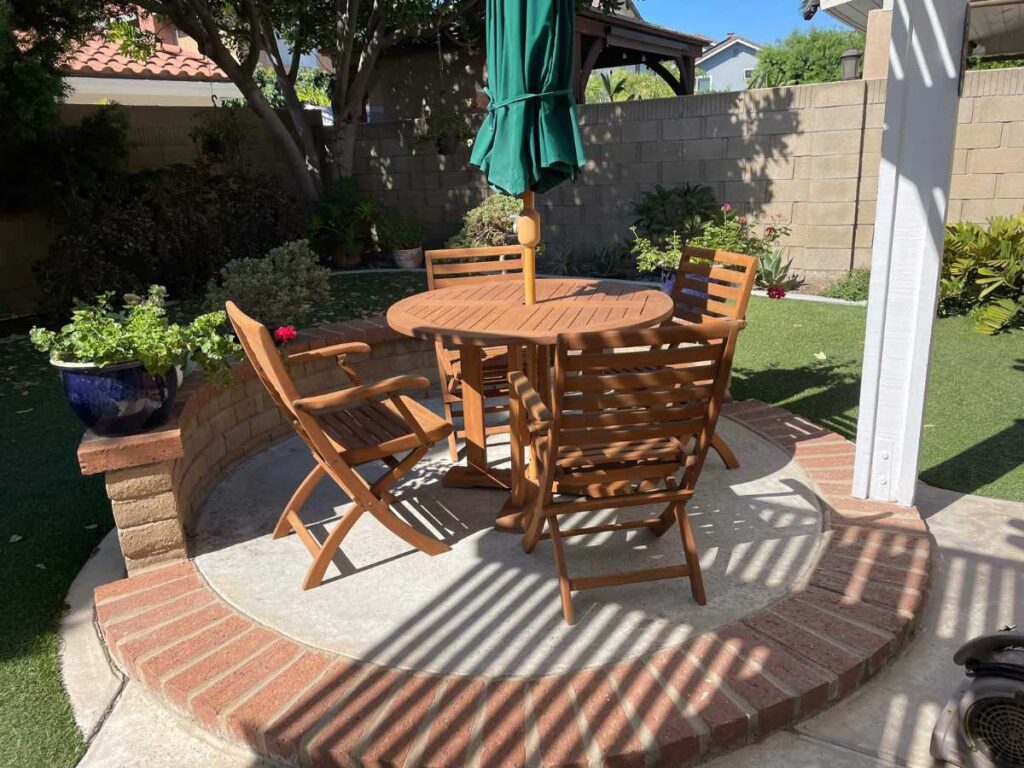 Sunlit backyard setting featuring a round wooden table surrounded by four folding chairs and a green patio umbrella.