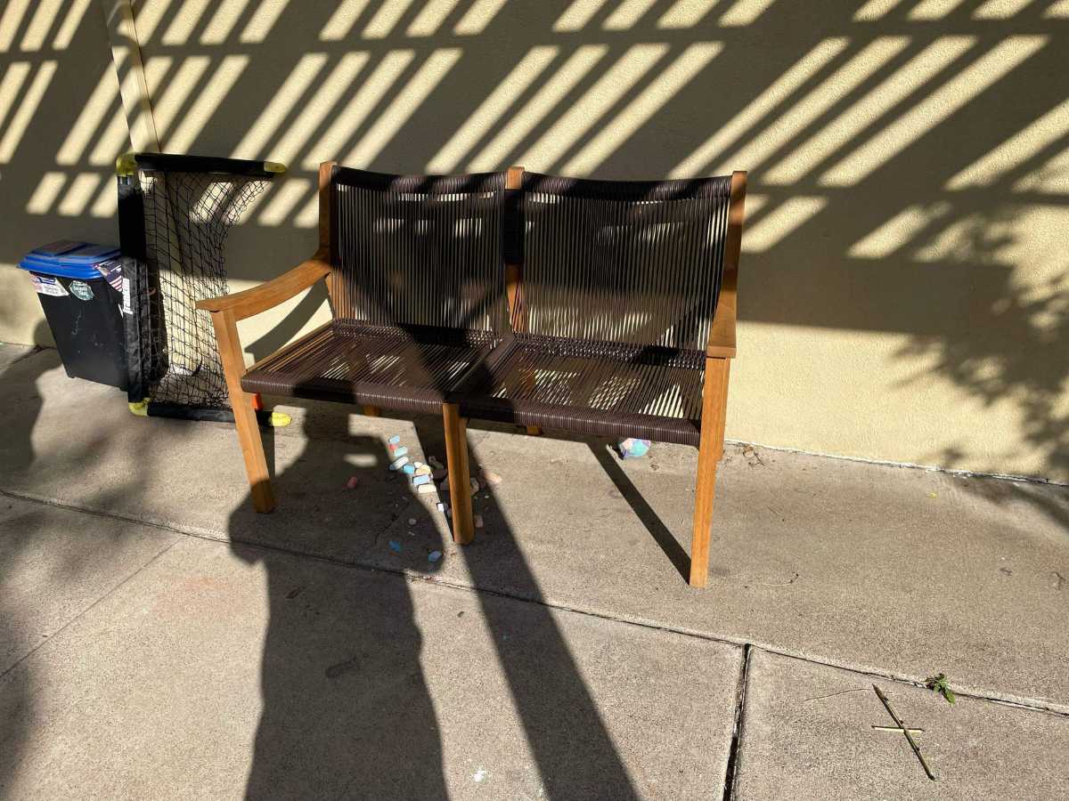 A two-seater wooden bench casts shadows on a wall beside a trash bin, with scattered colorful chalk pieces on the pavement.