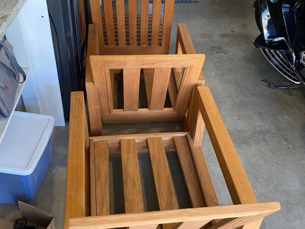 Two unfinished wooden chairs are positioned in a garage, alongside a motorcycle and storage containers.