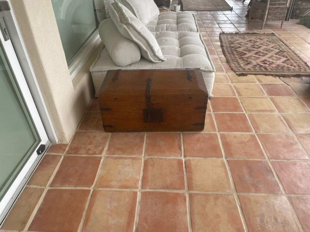 A rustic wooden storage trunk sits on a tiled patio beside a plush sofa, adding vintage charm to the outdoor space.