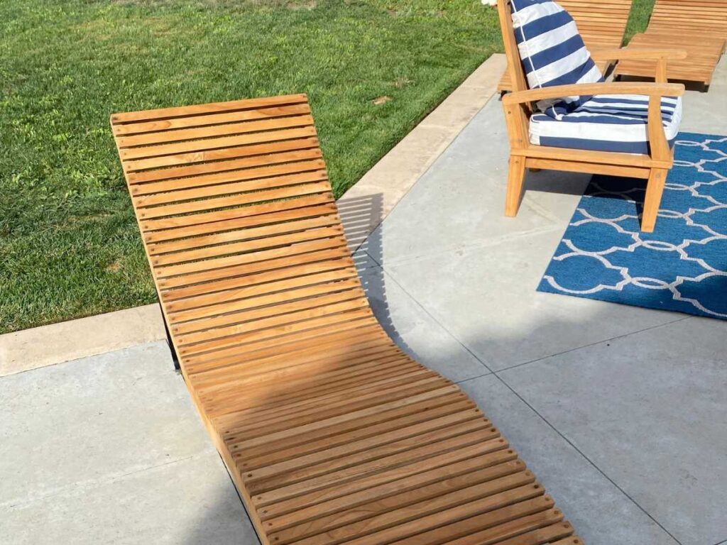 A wooden lounge chair with slatted design rests on a patio beside a green lawn and a blue patterned rug with a striped armchair.