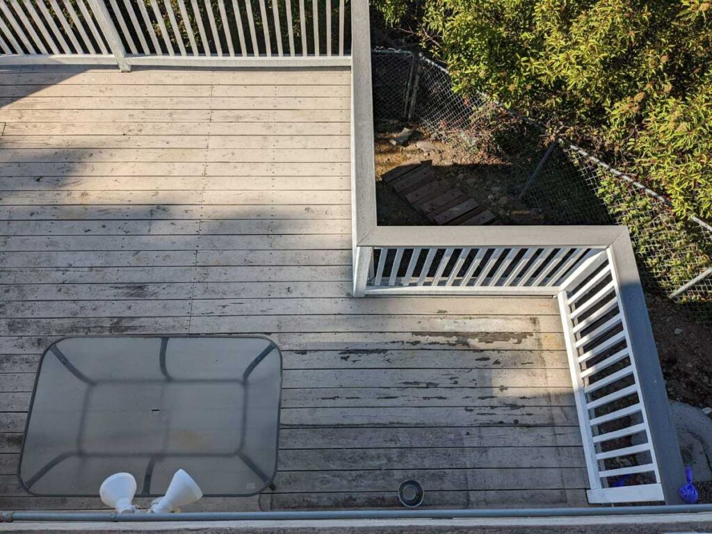 Aerial view of a wooden deck with a glass table, white railings, and a glimpse of shrubs and a fence in the background.
