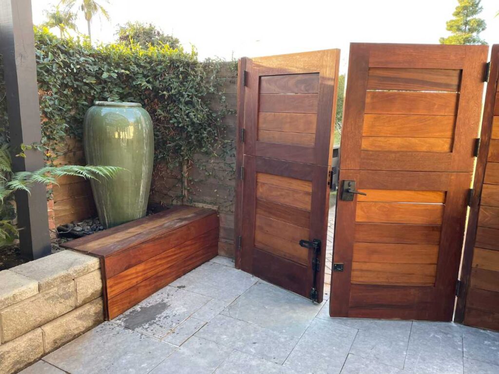 Two wooden gates open to a serene garden corner featuring a large green ceramic vase, a wooden bench, and lush greenery.