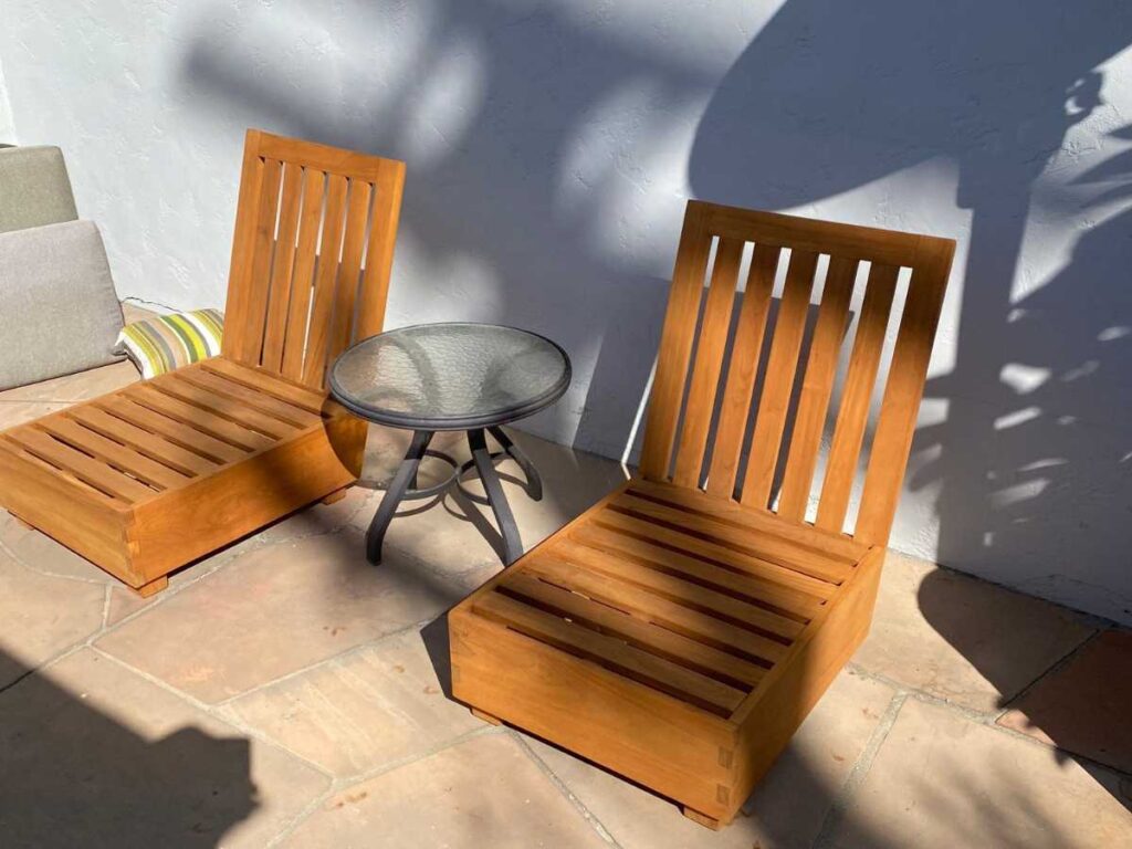 Two wooden lounge chairs sit beside a glass-topped table on a tiled patio, with shadows cast by a nearby wall.