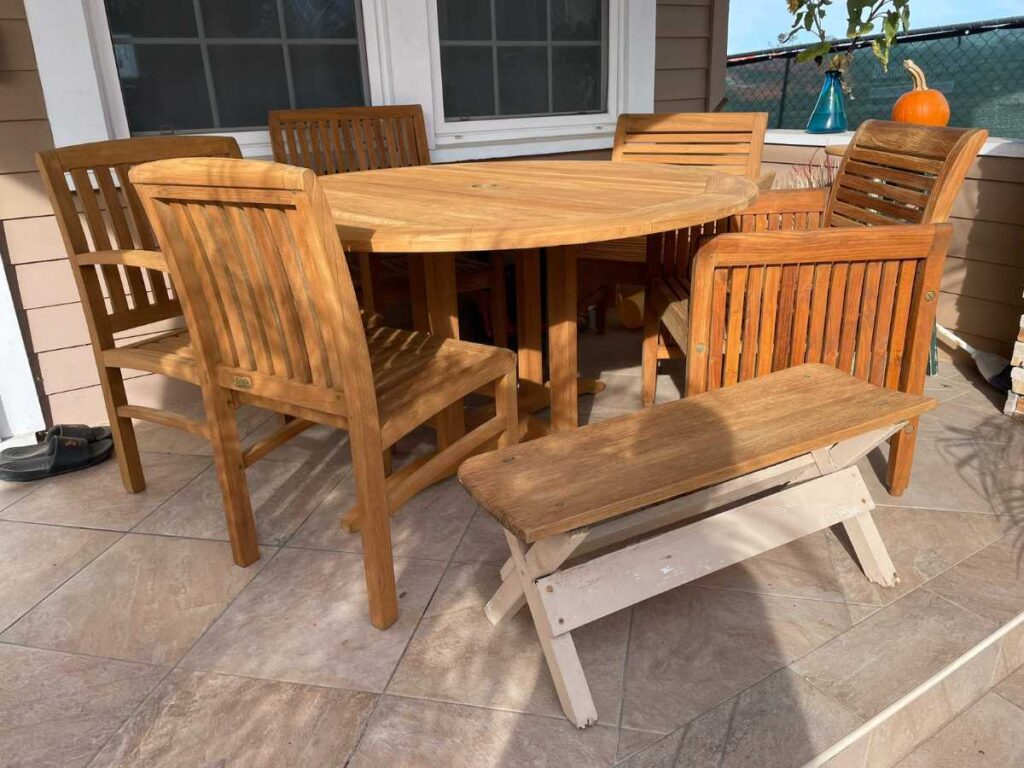 A wooden outdoor table surrounded by chairs, with a small white bench beside it, set on a tiled patio with sunlight shining.
