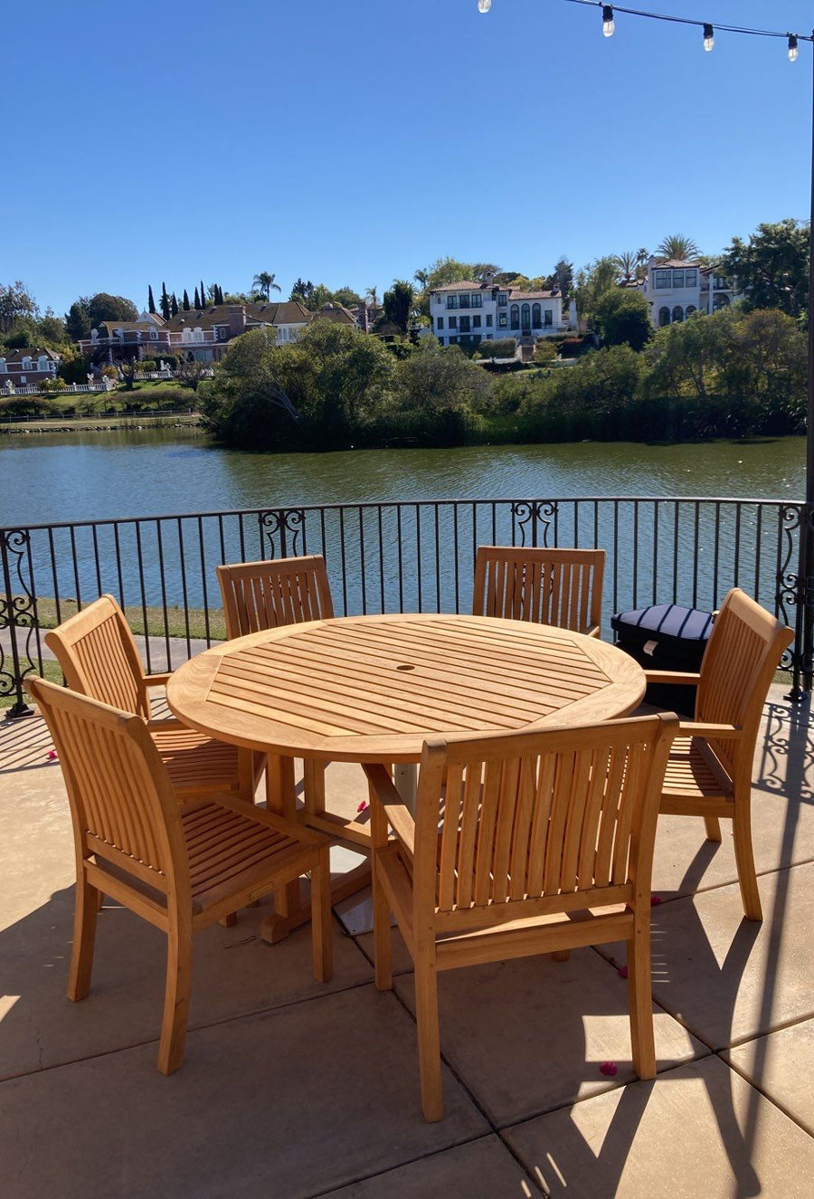 A wooden round table surrounded by chairs on a patio overlooking a serene lake and luxurious homes under a clear blue sky.