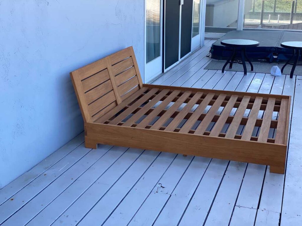 A wooden slatted bed frame rests on a white deck, leaning against a blue wall with sliding glass doors in the background.