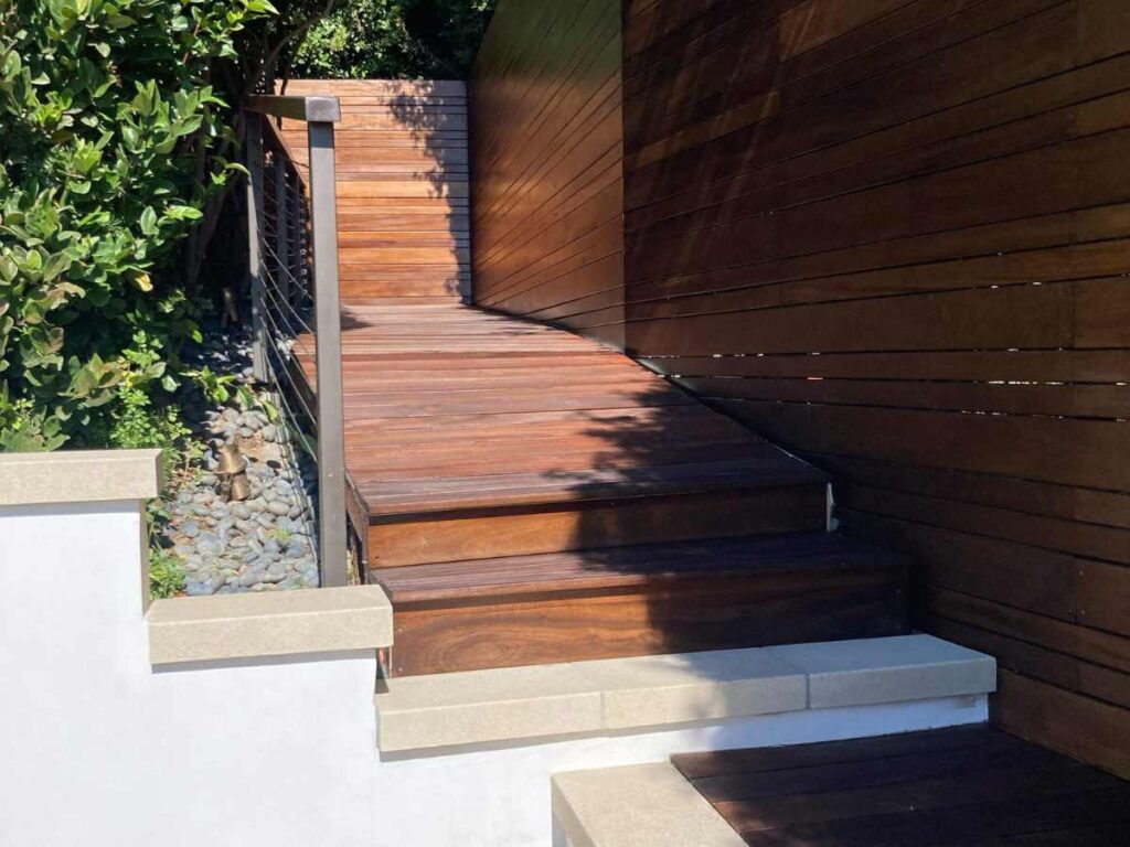 A wooden staircase leads up to a brown-paneled wall, surrounded by lush greenery and smooth stones. Sunlight casts gentle shadows.