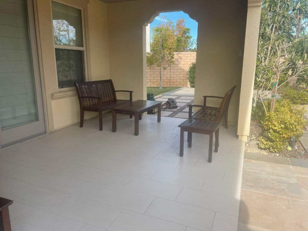 A covered patio with wooden chairs and a table, framed by a view of a landscaped garden and blue sky beyond.