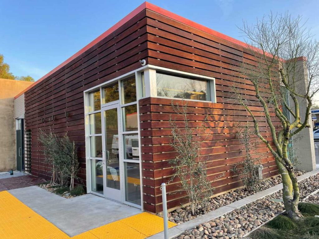 Modern building featuring wooden slat exterior, large glass doors, and landscaped pathway with rocks and small plants.