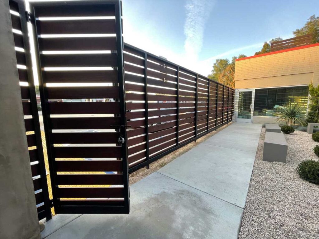 A tall, modern fence gate opens to a clean concrete pathway lined with decorative gravel and greenery, leading to a contemporary building.