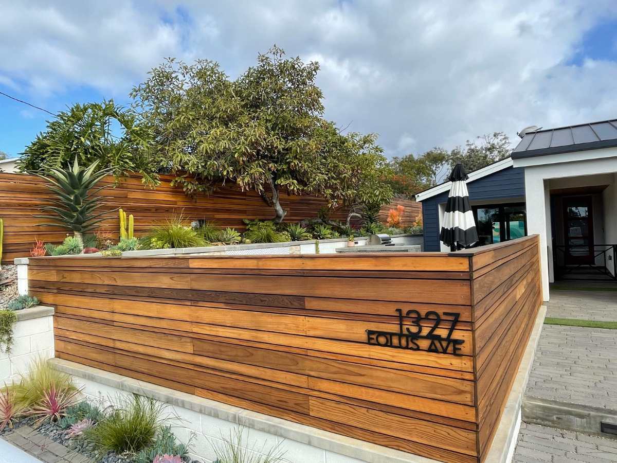 A modern home exterior featuring wooden fencing, lush greenery, succulents, and a black-and-white striped umbrella by the entrance.