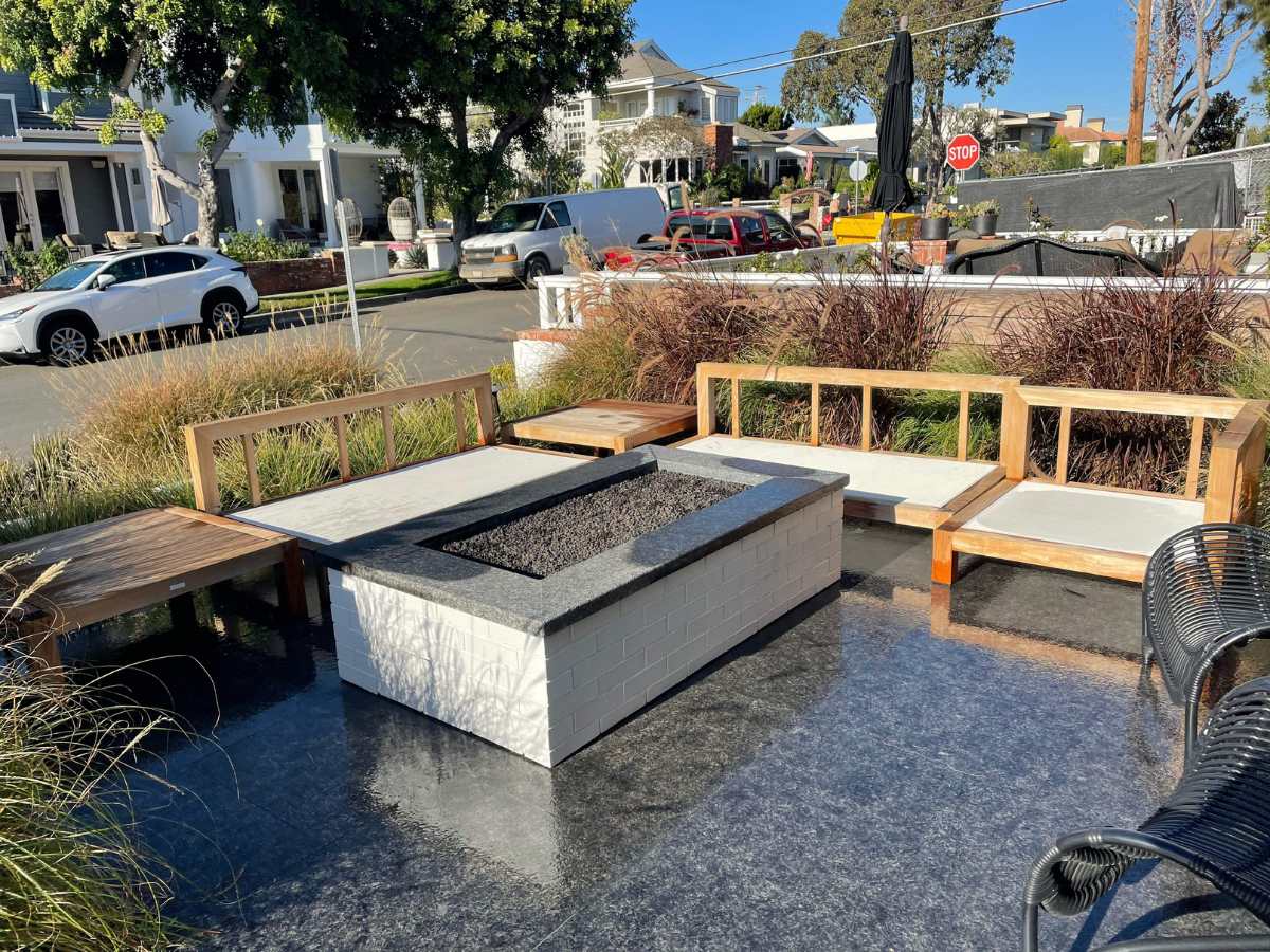 A modern outdoor seating area featuring wooden benches and a gas fire pit, surrounded by landscaping and parked cars nearby.