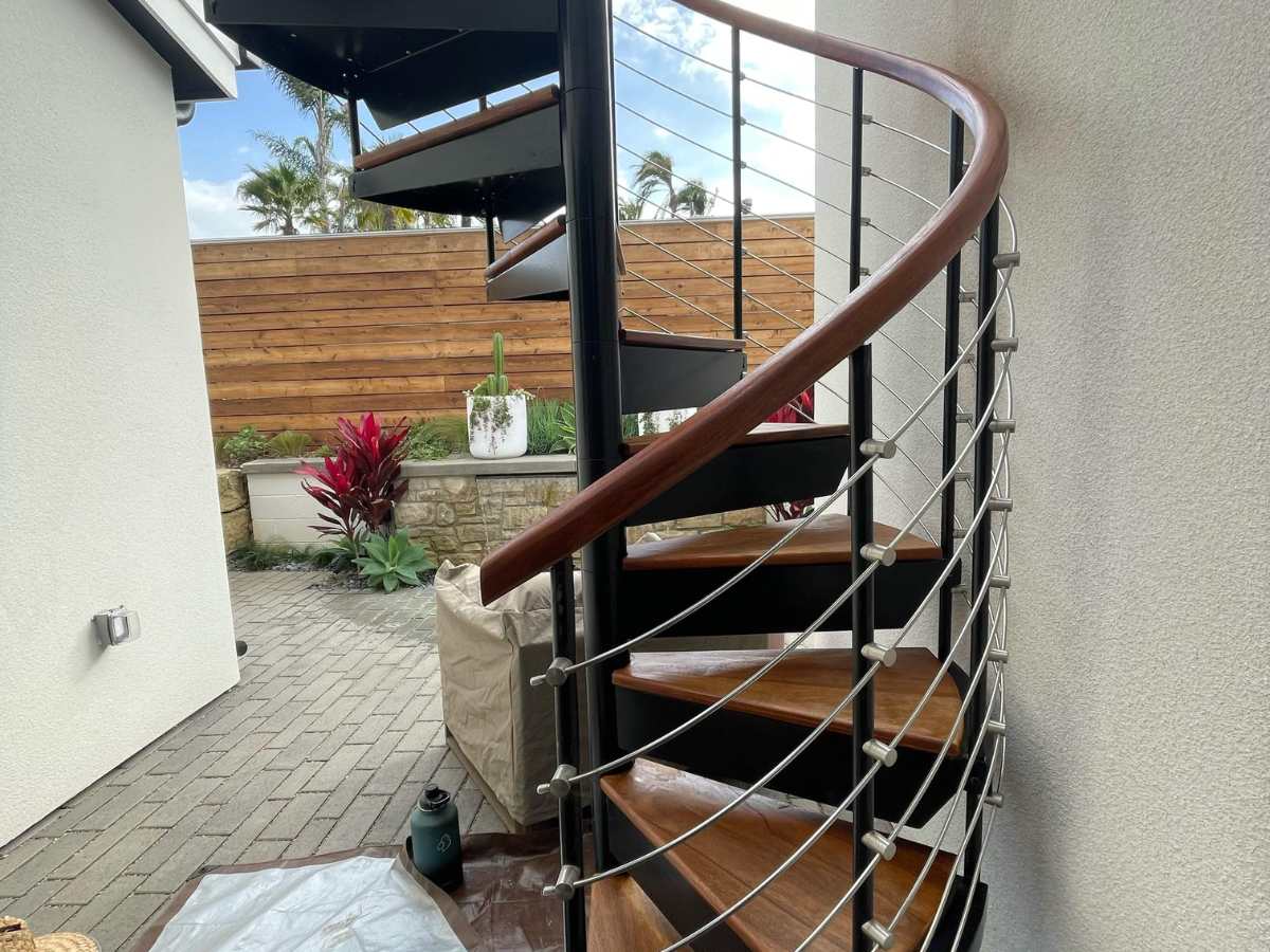 A modern spiral staircase with wooden steps and metal railing, set in a courtyard with tropical plants and a wooden fence in the background.
