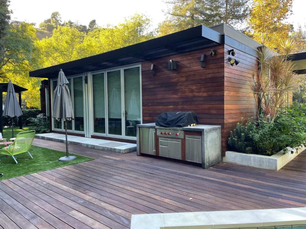 Modern wooden cabin with a patio, outdoor kitchen, and green garden, surrounded by trees and natural light.