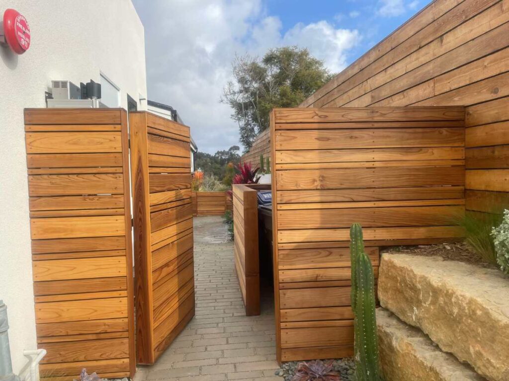A pathway lined with wooden fences leads to an outdoor space, featuring a cactus and stone steps against a cloudy sky backdrop.