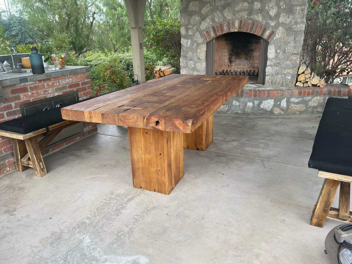 A rustic wooden dining table sits on a stone patio, accompanied by benches and a brick barbecue, surrounded by greenery.