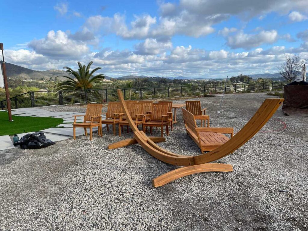 A serene outdoor space featuring wooden chairs, a curved lounge chair, gravel ground, and a palm tree against a cloudy sky.