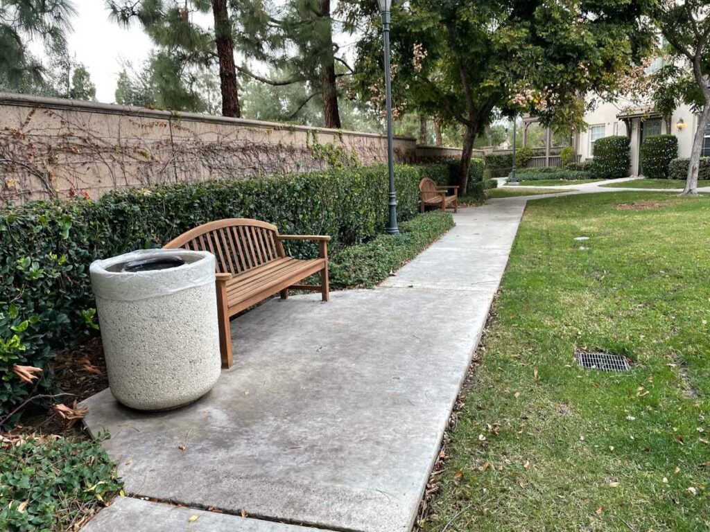 A serene walkway lined with benches and greenery, featuring a stone trash can amidst lush plants and trees.