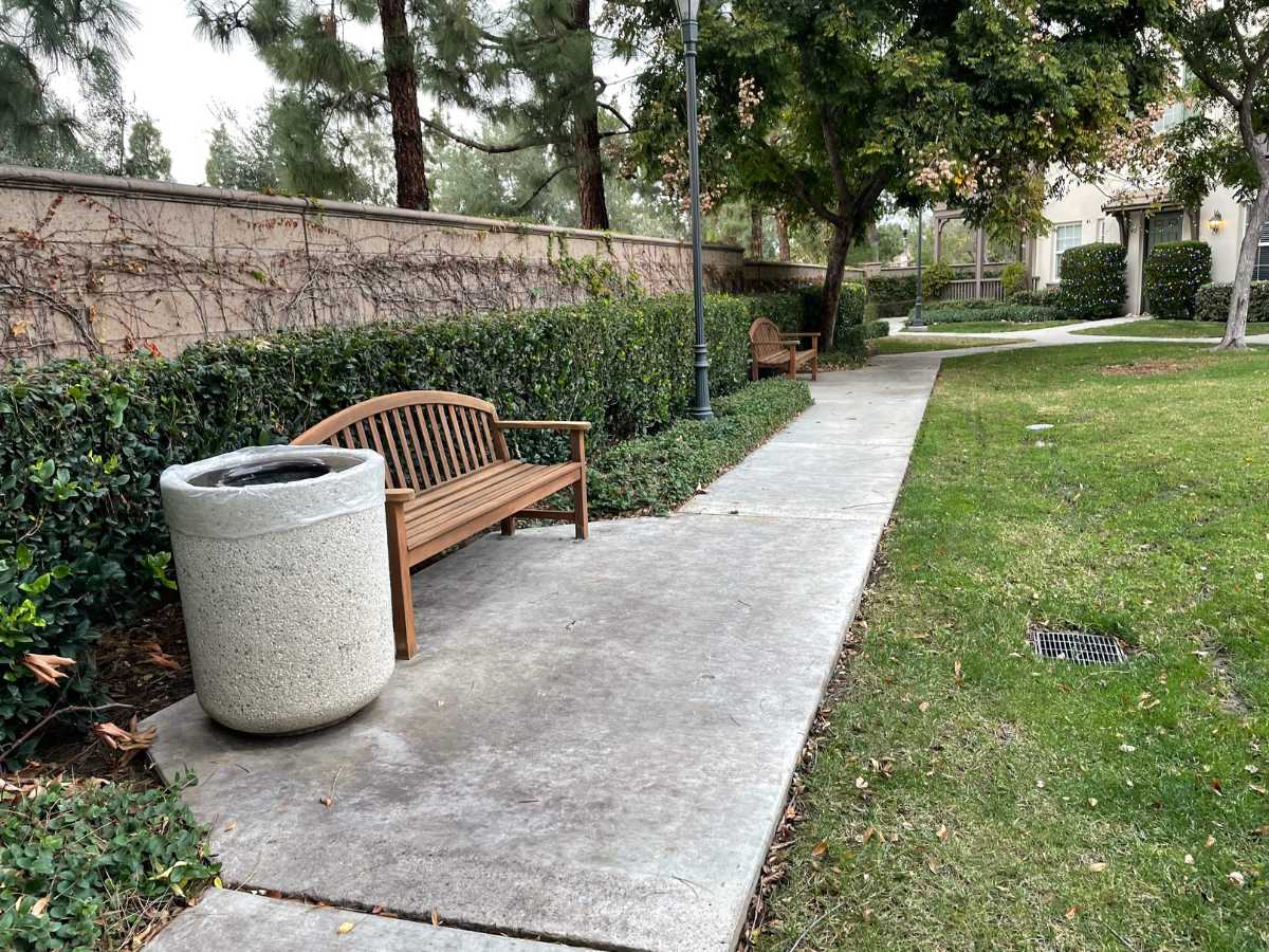 A serene walkway lined with benches and greenery, featuring a stone trash can amidst lush plants and trees.