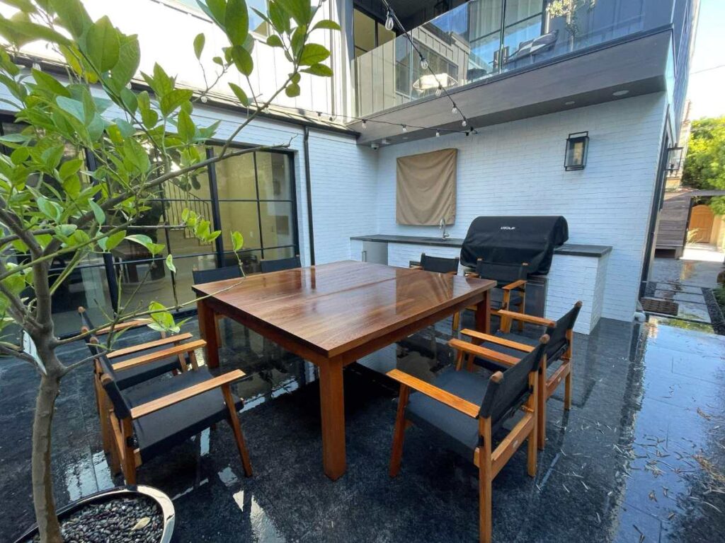 A stylish outdoor dining area features a wooden table, black chairs, and a grill against a modern home backdrop, surrounded by greenery.