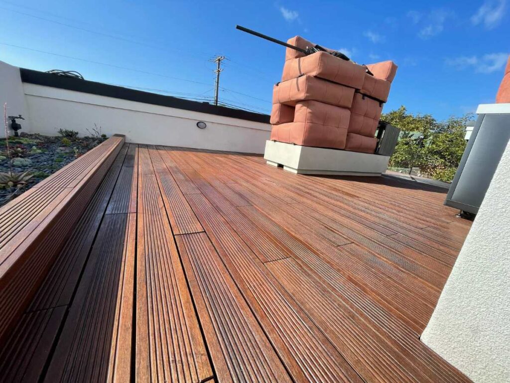 A sunlit wooden deck featuring stacked cushions, against a clear blue sky and greenery in the background.