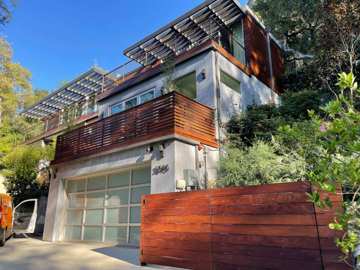 Modern two-story home with wooden slats, large windows, and a landscaped entrance, framed by greenery under a clear blue sky.