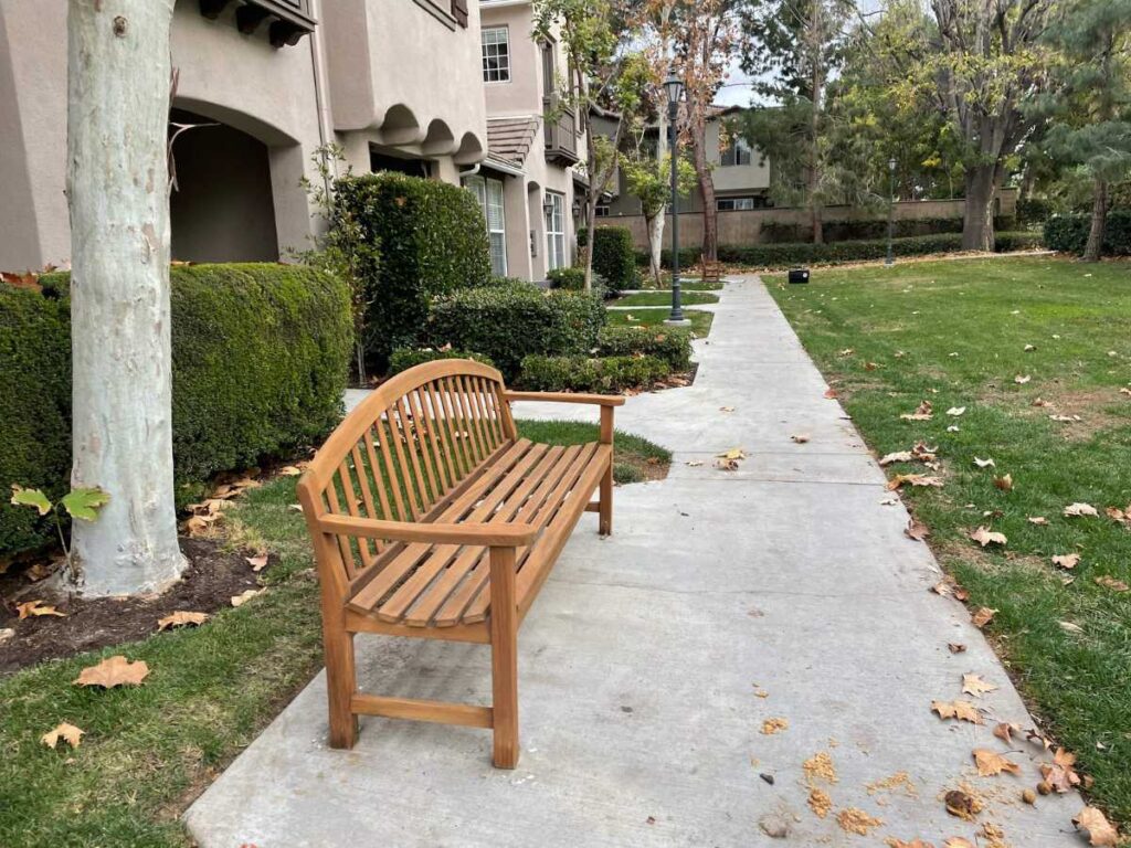 A wooden bench sits beside a paved walkway, surrounded by neatly trimmed hedges and trees in a serene, landscaped area.