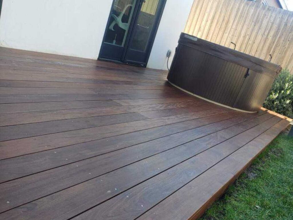 A wooden deck in a backyard featuring a hot tub on the right side, adjacent to a house with a sliding glass door.